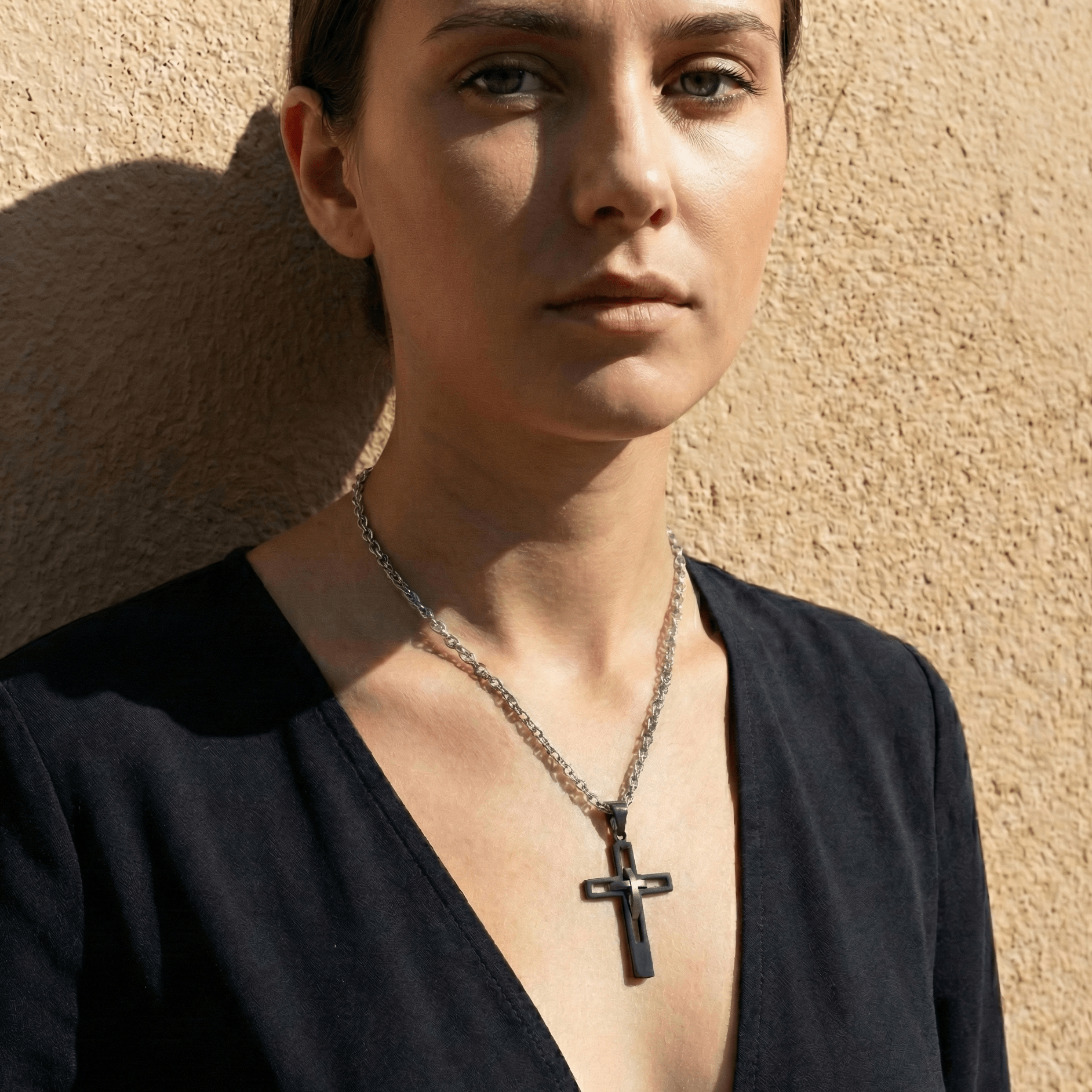 Woman wearing a black top and necklace with a cross pendant against a beige wall.